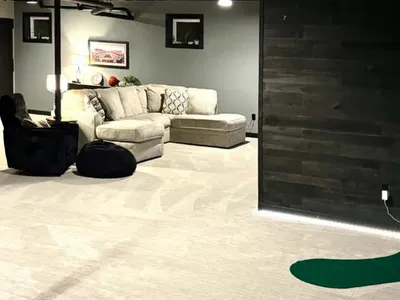 view from an open room with gray couch, black chair, and black wood paneling. A small putting green sits in the foreground